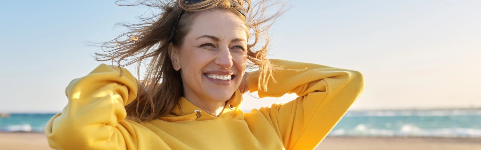 Eine selbstbewusste Frau um die 40 lächelt glücklich mit vom Wind verwehten Haaren am Strand.