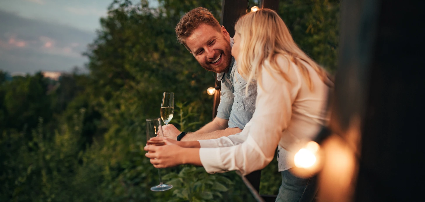 Ein glücklich lachendes Paar steht abends auf einem Balkon mit Lichterketten und stößt mit Sektgläsern an.