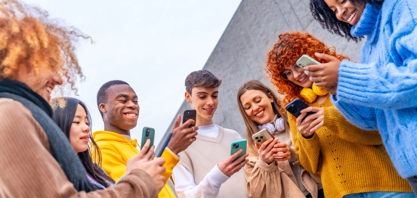 Eine Gruppe junger, diverser Menschen steht lachend im Kreis und nutzt verschiedene Smartphones, um neue Kontakte zu knüpfen.