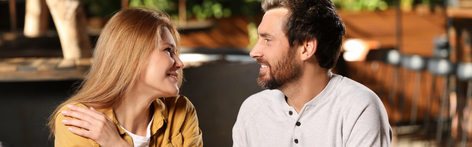 Ein Mann und eine Frau sitzen sich sympathisch lächelnd im Außenbereich eines Cafés gegenüber und unterhalten sich.