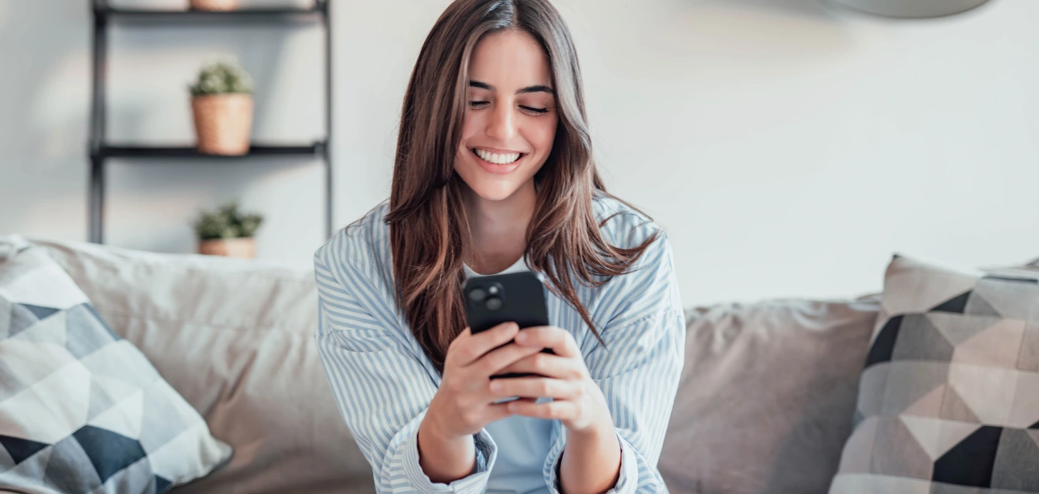 Eine lächelnde Single-Frau sitzt auf dem Sofa und chattet oder flirtet über ihr Smartphone.